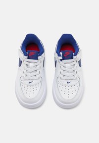White leather sneakers with blue accents and red interior. Features perforated toe, flat laces, and rubber sole. Nike logo on side.
