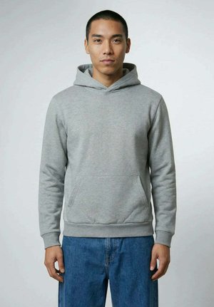 Junger Mann mit kurzen Haaren, der einen schlichten grauen Hoodie und blaue Jeans trägt, steht vor einem neutralen hellgrauen Hintergrund.