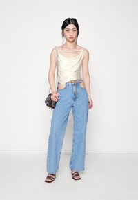 Camisola de cetim creme com alças finas, combinada com jeans de cintura alta azul claro com recortes laterais e um cinto de corrente dourada, complementada por saltos com tiras.