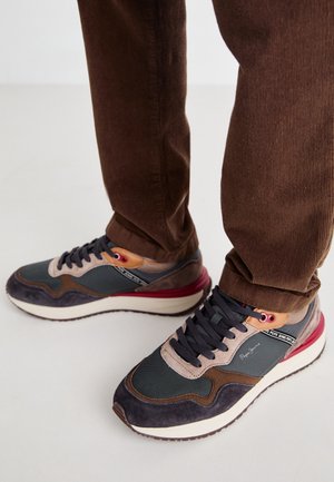 Person iført brune bukser og multifarvede sneakers med sorte snørebånd, beige og røde accenter på en lys gulvbaggrund.