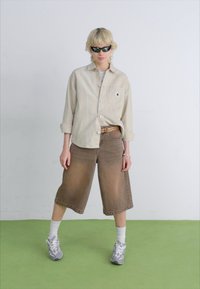Beige button-up shirt, brown denim culottes, gray athletic shoes, white crew socks, and black sunglasses. Minimalist design on a green floor.
