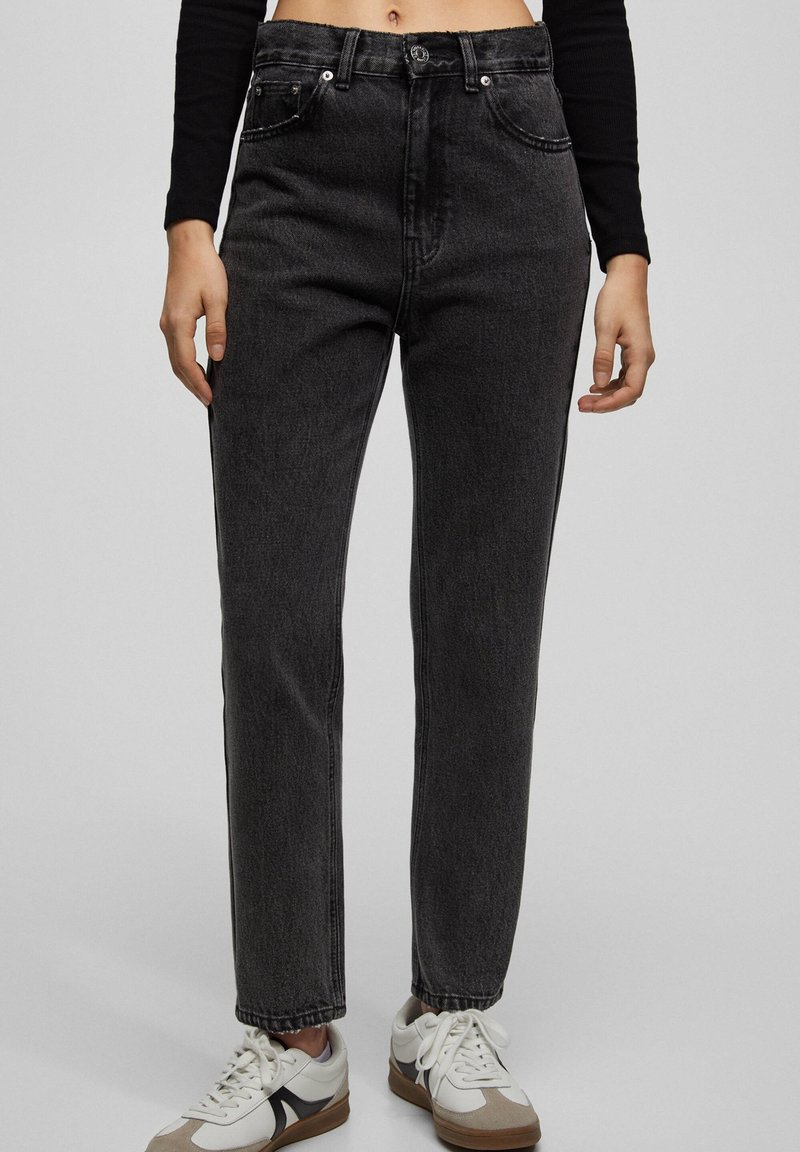 Jean taille haute, coupe droite en denim gris foncé avec une finition texturée, dotés de poches avant et d'une fermeture à bouton, associés à des baskets blanches.