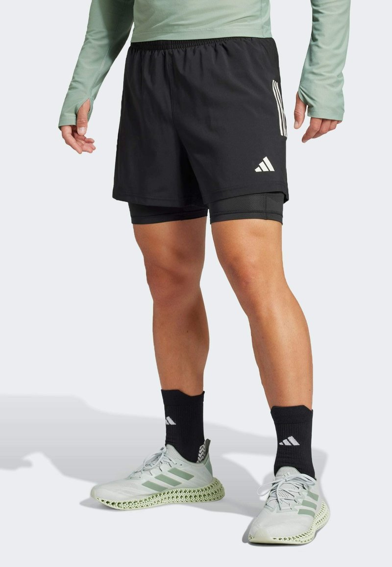 Personne portant un short de sport noir avec des shorts de compression intégrés, des chaussettes noires et des chaussures de course Adidas vert clair, debout sur un sol blanc.