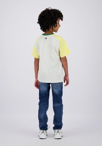 T-shirt grigia con maniche gialle corte, dettagli verdi e un'etichetta sul retro. Abbinato a jeans blu e sneakers bianche.