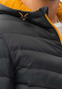 La chaqueta acolchada presenta un exterior de color gris oscuro con un forro de capucha amarillo brillante. Incluye un cierre frontal y un cordón ajustable para un ajuste ceñido.