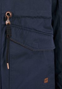 Navy jacket featuring a large front pocket, textured fabric, contrasting copper accents, and a drawstring with a metal toggle.
