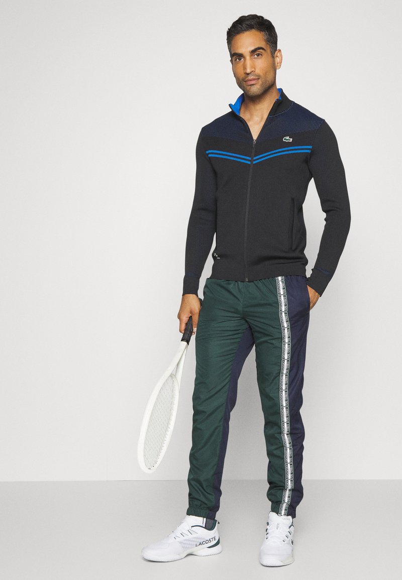 Veste zippée noire avec des rayures bleues et une texture tricotée, assortie à un pantalon de survêtement vert et marine avec des rayures sur les côtés, et des baskets blanches.