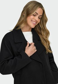 Cappotto in lana nero con colletto a scissione, caratterizzato da un design doppiopetto, tessuto testurizzato e due tasche frontali.