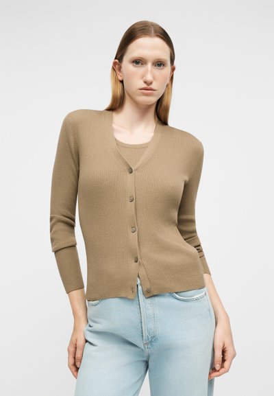 Γυναίκα που φοράει μπεζ ζέρσεϊ ζακέτα με κουμπιά πάνω από ένα αντίστοιχο τοπ και ανοιχτόχρωμα τζιν ψηλόμεσα, στέκεται μπροστά σε απλό φόντο.