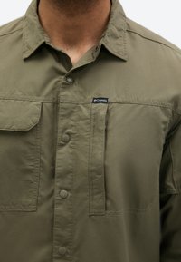 Közeli kép egy személyről, aki oliva zöld Columbia inget visel patentgombokkal és mellzsebekkel, a gallér látható, de az arc nincs a képen.