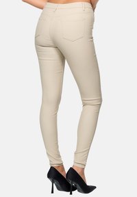 Jean skinny beige avec une texture lisse, taille mi-haute, deux poches arrière et une silhouette ajustée, accompagnés de chaussures à talons hauts noires.