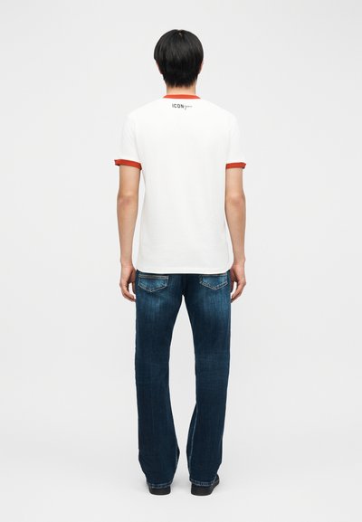 Άνδρας που στέκεται και κοιτάζει προς τα πίσω, φοράει λευκό μπλουζάκι με κόκκινες λεπτομέρειες και σκούρο μπλε τζιν, μπροστά από απλό φόντο.