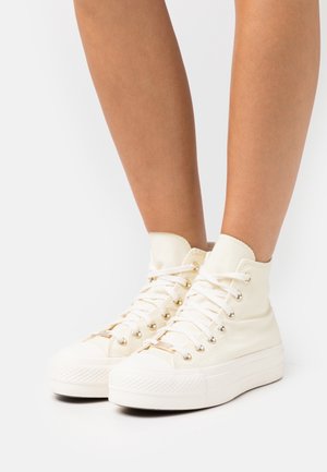 Sneakers alti in tela bianca con occhielli in tono oro, lacci piatti e suole in gomma testurizzata. Altezza alla caviglia con punta liscia e arrotondata.