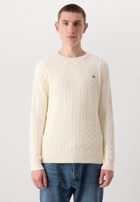 Maglione crema a trecce con scollo rotondo e polsini a coste, abbinato a jeans blu. Presenta un piccolo logo sul petto.