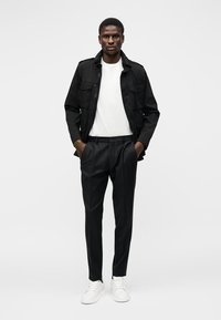 Άνδρας που φοράει μαύρο μπουφάν, λευκό μπλουζάκι, μαύρο παντελόνι και λευκά αθλητικά παπούτσια, στέκεται με τα χέρια στις τσέπες μπροστά σε απλό φόντο.