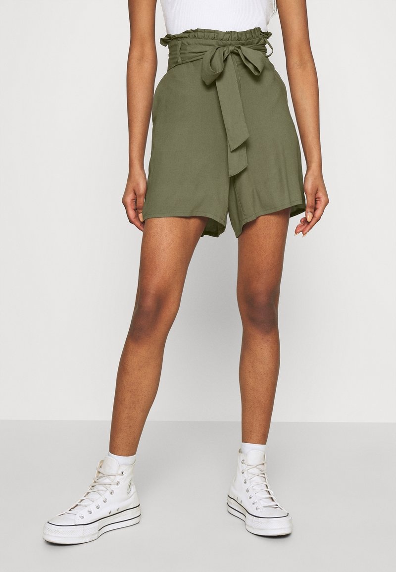 Olive green high-waisted shorts with a ruffled waistband and a tied bow, featuring a loose fit and rolled hem, paired with white high-top sneakers.