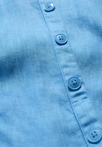 Svetle modrá lněná košile s šesti odpovídajícími knoflíky, jemnou texturou a zapínáním na knoflíky s čistým zakončením na okrajích.