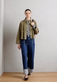 Olivgrön kort jacka över en svart och vit randig skjorta, tillsammans med högmidjade blå denimjeans med grå muddar och svarta skor.