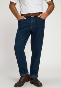 Jeans de mezclilla azul oscuro con un corte relajado, que presentan un cinturón marrón, detalles de costura sutiles y dobladillos enrollados sobre botas de cuero marrón.