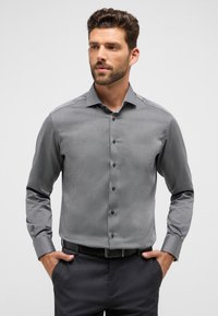 Graues Hemd mit Knöpfen, einem dezenten geometrischen Muster, langen Ärmeln und einem Kentkragen. Kombiniert mit dunklen Hosen und einem schwarzen Gürtel.