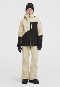 Ski-jack in beige en zwart met een geometrisch patroon, gecombineerd met bijpassende beige skibroek. Inclusief handschoenen en skibril.