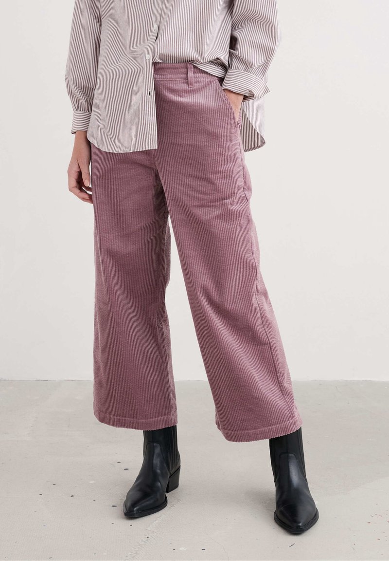 Personne portant un pantalon en velours côtelé mauve à jambes larges, une chemise rayée boutonnée et des bottines noires, debout sur un sol clair.