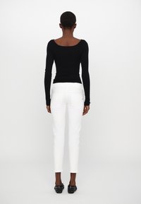 Kvinna som står vänd bort, iklädd en svart långärmad topp, vita slim-fit byxor och svarta nitar skor på en vit bakgrund.