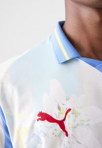 Polo majica kratkih rukava u svijetlo plavoj boji s cvjetnim uzorkom. Crveni Puma logo na prsima, plavi ovratnik s žutim akcentnim prugama. Tekstuirana tkanina.