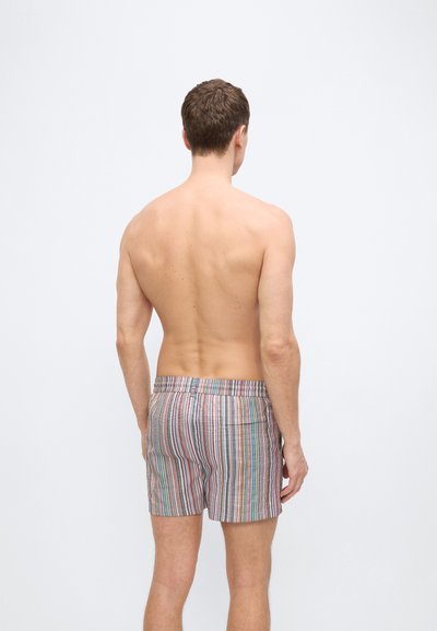 Άνδρας που στέκεται γυρισμένος, χωρίς πουκάμισο, φοράει πολύχρωμο κοντό παντελονάκι με κάθετες ρίγες μπροστά σε απλό ανοιχτό φόντο.