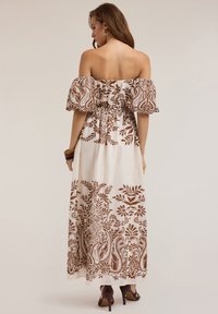 Robe maxi décolletée en tissu blanc avec un imprimé floral et paisley marron, taille froncée et manches bouffantes. Portée avec des sandales à talons.