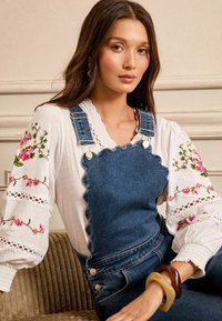 Denim salopette s špageti robovi, srebrnimi detajli in nastavljivimi naramnicami, nosene preko bele bluze z rožnatimi cvetličnimi vezeninami in čipkastimi detajli.