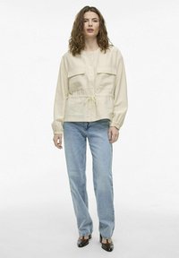 Beige bunda s gombíkmi a dvoma prednými vreckami, elastickým pásom a sťahovacou šnúrkou. Skombinovaná s svetlomodrými džínsami a hnedými podpätkami.