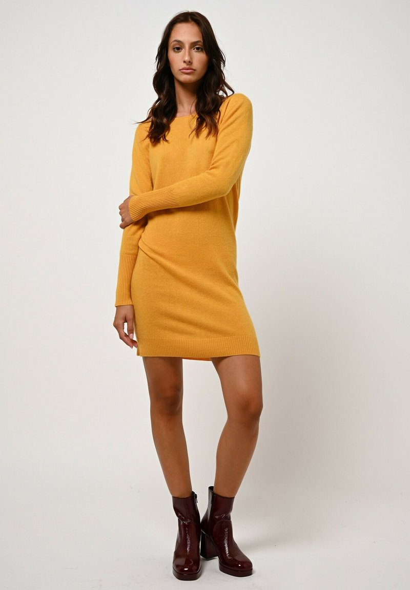 Donna in piedi che indossa un vestito a maglia giallo senape a maniche lunghe e stivaletti rossi scuri fino alla caviglia, su uno sfondo bianco semplice.