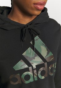 Hoodie noir en coton avec un logo Adidas au motif camouflage. Il possède une capuche avec cordon de serrage et une impression texturée. Gros plan sur l'encolure.