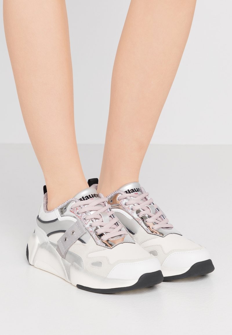 Zapatillas en gris claro y blanco con acentos metálicos, tejido texturizado, cordones rosas y suela de goma negra. Presentan un logo en la lengüeta.
