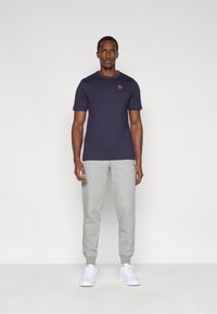 Μπλε μαρινιέρα κοντομάνικη μπλούζα με στρογγυλή λαιμόκοψη και μικρό λογότυπο στο στήθος, συνδυασμένη με ανοιχτό γκρι φόρμα και λευκά αθλητικά παπούτσια.