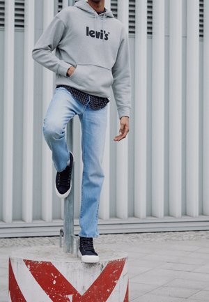 Personne portant un sweat à capuche gris Levi's, jean bleu clair, baskets noires, appuyée avec un pied sur un bloc de béton rayé rouge et blanc en plein air.