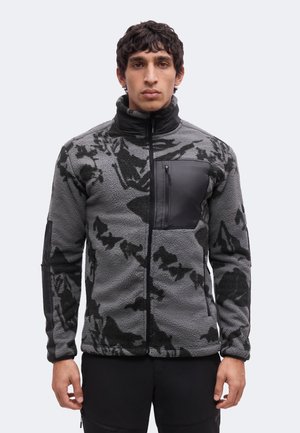 Άνδρας που φορά ένα γκρι και μαύρο fleece με σχέδιο, με ψηλό γιακά και τσέπη στο στήθος με φερμουάρ, στέκεται μπροστά σε απλό φωτεινό φόντο.