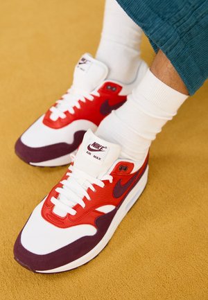Personne portant des baskets Nike Air Max blanches, rouges et violet foncé avec des chaussettes blanches et un pantalon bleu sarcelle sur un tapis jaune.
