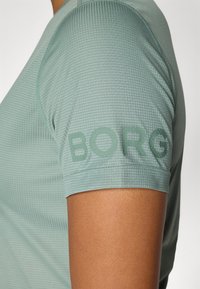 Svetlo zelena atletska majica iz teksturiranega blaga, s kratkimi rokavi in napisom "BORG" natisnjenim v temnejši zeleni barvi na desnem rokavu.