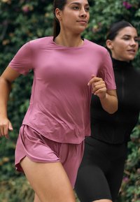 Růžový sportovní top s krátkým rukávem a odpovídající šortky z lehkého texturovaného materiálu. Dva běžci v pohybu, v pozadí zeleň.