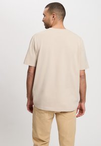 Beżowa koszulka z krótkim rękawem o luźnym kroju, gładkiej fakturze i okrągłym dekolcie, połączona z jasnobeżowymi spodniami wyposażonymi w tylne kieszenie.