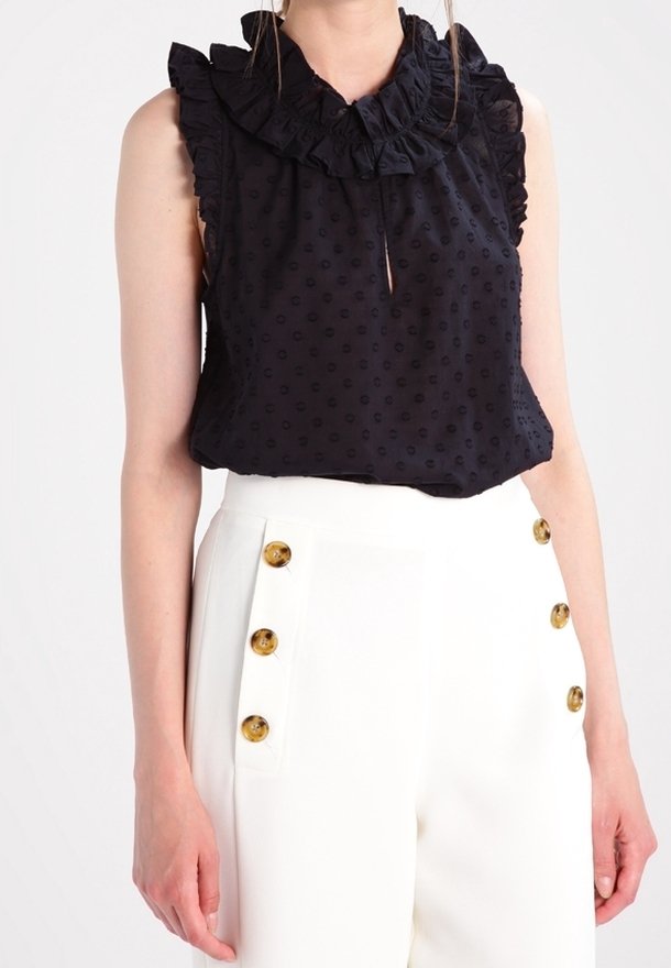 Blouse noire sans manches avec col volanté et pois texturés, associée à un pantalon blanc taille haute à deux rangées de boutons marron.