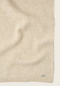 Couverture en tricot beige avec un motif en rib texture, présentant une petite étiquette de marque métallique dans le coin inférieur. Tissu doux et chaud.