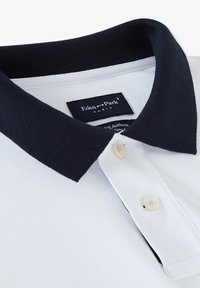 Polo blanc avec un col bleu marine. Comprend deux boutons et un tissu texturé. L'étiquette indique "Eden Park Paris." 100 % coton.