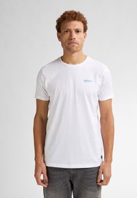 T-shirt bianco in cotone con maniche corte, colletto tondo e piccolo dettaglio in testo blu sul petto. Indossato con pantaloni in denim grigi.