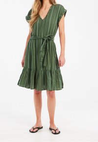 Grünes Kleid mit vertikalen Streifen und kleinen weißen Punkten. V-Ausschnitt, kurze Ärmel, tailliert mit Gürtel und einem gerafften Saum. Getragen mit schwarzen Sandalen.