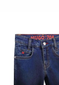 Jeans de mezclilla en azul oscuro, con una cinturilla de marca roja, botón de metal y detalles de costura, además de una pequeña etiqueta roja en el bolsillo delantero.