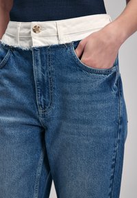 Jeans de denim en azul medio con una cinturilla blanca texturizada. Cuenta con un bolsillo frontal y un botón metálico visible. Detalle de dobladillo desgastado.