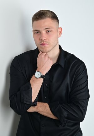 Schwarzes Langarmshirt, getragen von einer Person mit kurzen Haaren, die eine silberne Armbanduhr mit weißem Ziffernblatt und schwarzem Armband trägt. Der Hintergrund ist schlicht weiß.